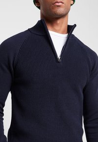 Granatowy sweter zapinany na suwak wykonany z prążkowanego dzianinowego materiału, z golfem, srebrnym zamkiem błyskawicznym i kontrastową fakturą na rękawach.