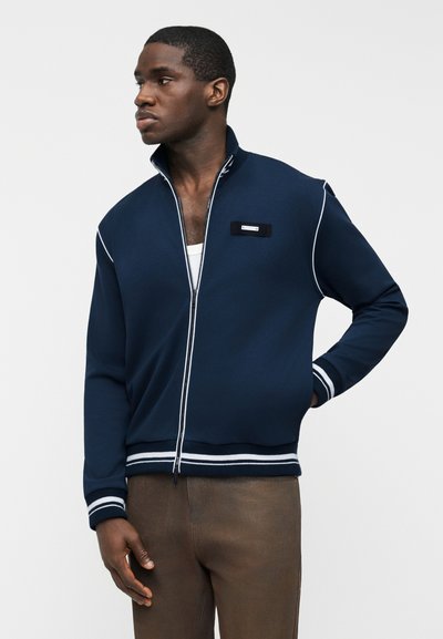 Homme portant une veste zippée bleu marine avec bordure blanche et un pantalon marron, regardant vers la gauche avec une main dans la poche.