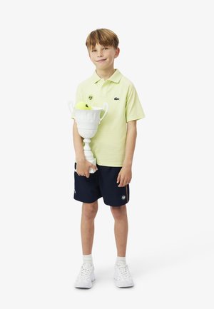 Ragazzo con polo gialla e pantaloncini blu scuro che tiene un trofeo da tennis bianco con una pallina da tennis gialla all'interno, in piedi su sfondo bianco.