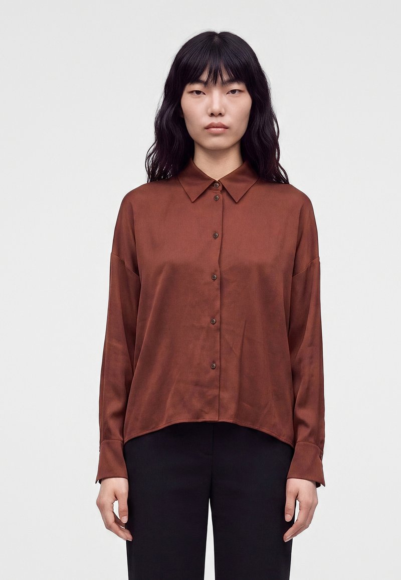 Femme aux longs cheveux noirs portant une chemise marron boutonnée et un pantalon noir, debout devant un fond clair uni.