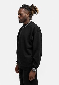 Schwarzer Baumwollpullover mit Rundhalsausschnitt und gerippten Bündchen. Das Modell trägt passende schwarze Jogginghosen und ein silbernes Kettenarmband.