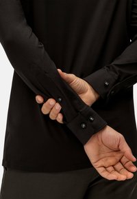 Zwarte lange mouwen shirt met knoopmanchetten, slanke stof textuur en een nauwsluitend ontwerp, gecombineerd met donkere broek.