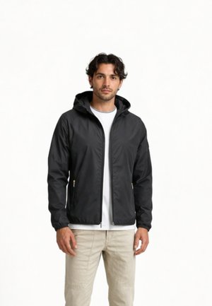 Uomo che indossa una giacca nera con cappuccio e cerniera sopra una maglietta bianca e pantaloni beige, in piedi davanti a uno sfondo bianco semplice.