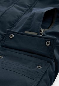 Giacca blu navy con texture trapuntata, dotata di una tasca con zip e dettagli a pressione, realizzata in materiale liscio e resistente alle intemperie.