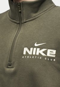 Κοντινό πλάνο σε ελιά πράσινη φόρμα με φερμουάρ μέχρι το λαιμό, με λευκό κεντητό λογότυπο Nike και το κείμενο «Athletic Club» στο στήθος.