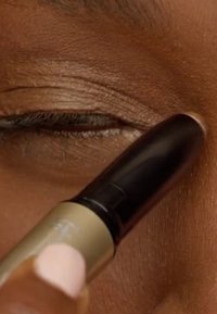 Aplicando corrector beige en el párpado cerrado con el dedo, sujetando el lápiz de maquillaje cerca de la esquina externa del ojo.