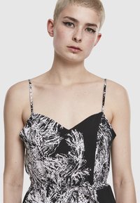 Persoană cu păr scurt blond, purtând o rochie cu bretele subțiri, cu un model abstract alb-negru, expresie neutră și piercinguri faciale.