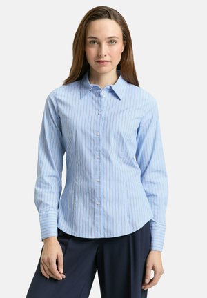 Mujer con una camisa de manga larga color azul claro con botones y finas rayas verticales, combinada con pantalones azul marino oscuro sobre un fondo blanco.