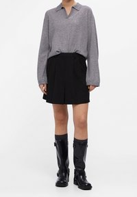 Polo gris en tricot, jupe mini plissée noire et bottes noires brillantes jusqu'aux genoux avec des boucles. L'ensemble présente une coupe décontractée et des textures superposées.