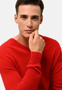 Roter gestrickter Pullover mit V-Ausschnitt und gerippten Bündchen, der eine glatte Textur und einen eng anliegenden Stil aufweist, getragen von einem Modell mit der Hand am Kinn.