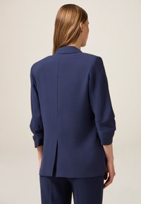 Blazer blu navy su misura con una fessura singola, maniche su misura arrotolate ai polsi, realizzato in tessuto liscio, caratterizzato da un design minimalista.