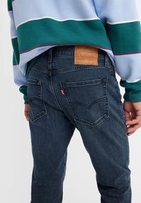 Personne portant un jean Levi's bleu foncé et un pull rayé bleu, vert et blanc, montrant les poches arrière et le patch en cuir de la marque.