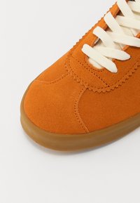 Gros plan sur l'avant d'une chaussure de sport en daim orange avec des lacets blancs et une semelle en caoutchouc marron sur un fond clair.