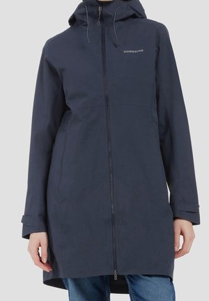 Femme portant une veste imperméable à capuche bleu marine foncé avec une fermeture éclair frontale et le logo Didriksons sur la poitrine.