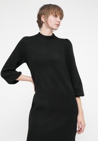 Melna adīta kleita ar augstu kakla izgriezumu, 3/4 garuma piedurknēm un ribotu apakšmalu. Audums izskatās mīksts ar gludu tekstūru.