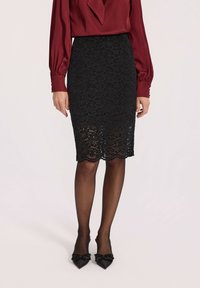 Kobieta w czarne koronkowej spódnicy ołówkowej, przejrzystych czarnych rajstopach, czarnych szpiczastych szpilkach z kokardkami oraz długim rękawie bordowej bluzce z guzikami przy mankietach.