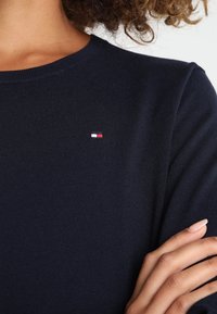 Närbild av en person iförd en marinblå stickad tröja med en liten rektangulär logotyp i rött, vitt och blått på bröstet.