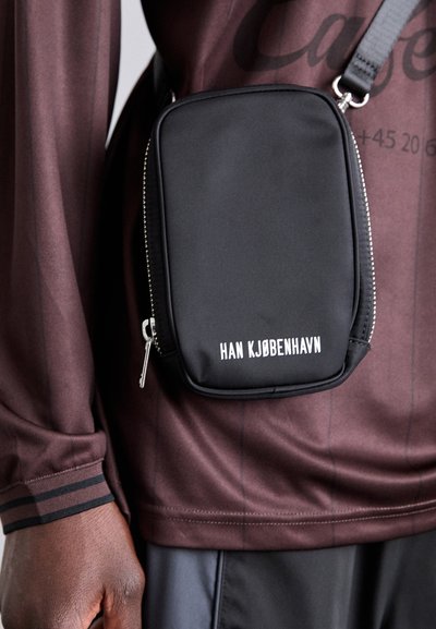 Black fabric crossbody bag with a rectangular shape, zip closure, and white text "HAN KJØBENHAVN" on the front. Matte finish and minimal design.