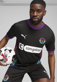 Camiseta de fútbol negra con cuello y mangas moradas, que presenta un patrón de degradado colorido en los laterales y un logo de patrocinador blanco. Sosteniendo un balón de fútbol.