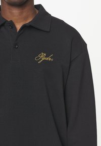 Černé polo tričko vyrobené z bavlny, s límečkem a dvěma knoflíky. Zlatě vyšívané logo na levém prsu. Hladká textura.