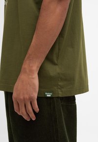 T-shirt en coton vert olive avec un col rond, doté de manches courtes et d'une étiquette sur le côté avec le logo 'Barbour'.