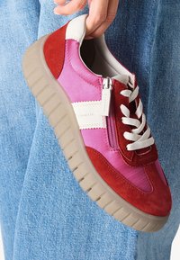 Les sneakers présentent un mélange de daim rouge et rose avec des accents blancs, une fermeture éclair sur le côté et une épaisse semelle texturée beige. Les lacets sont blancs.
