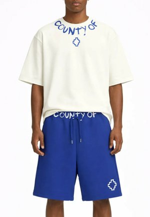 Uomo che indossa una maglietta bianca oversize con la scritta "COUNTY OF" e pantaloncini blu con coulisse e logo geometrico sul davanti.
