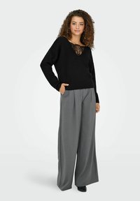 Zwarte trui met een V-hals en kantaccent, gecombineerd met hooggeviseerde, wijde grijze broeken. Soepele textuur, relaxed fit.