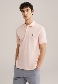 Svetloružová polo tričko s krátkymi rukávmi, golierom, trojriadkovým zapínaním na gombíky a malým čiernym logom. Nosené s svetlosivými textúrovanými nohavicami.