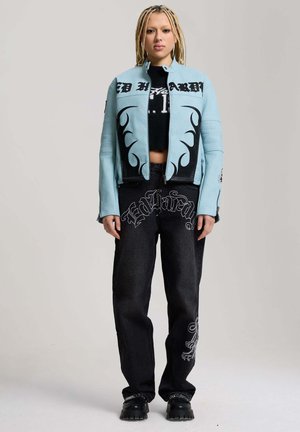Osoba z długimi zaplecionymi włosami, ubrana w jasnoniebieską kurtkę z czarnymi wzorami płomieni oraz czarne spodnie z dekoracyjnym białym tekstem i wzorami.