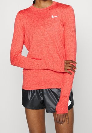 Personne portant un haut de sport Nike corail à manches longues avec des trous pour les pouces et un short noir Nike, debout avec un bras croisé sur l'autre.