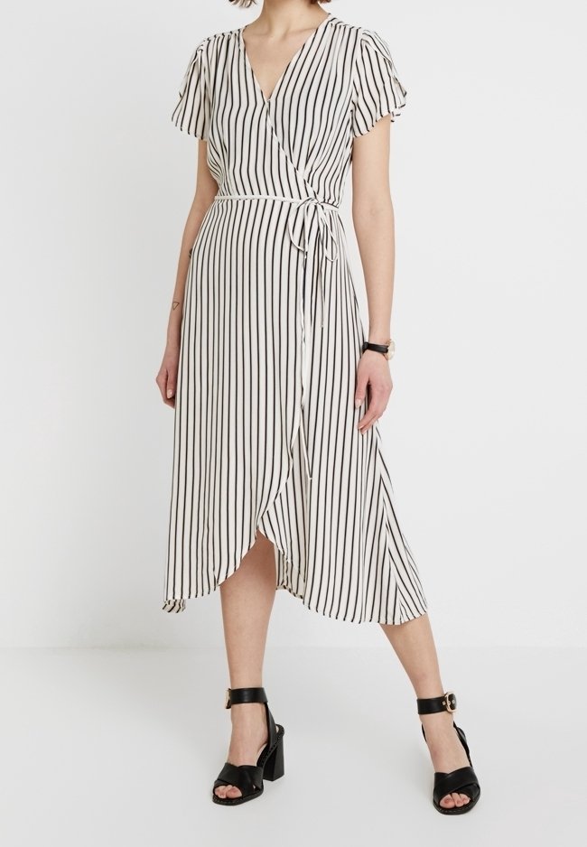 Femme portant une robe portefeuille midi blanche à rayures verticales noires et des sandales à talons carrés avec bride à la cheville noire, debout devant un fond uni.