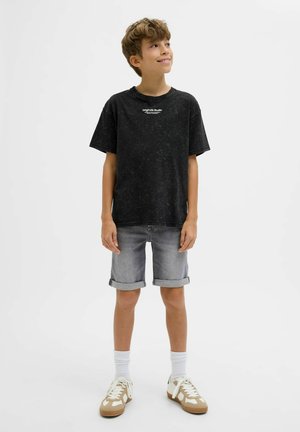 Zwarte t-shirt met korte mouwen en een gestructureerde afwerking, witte tekst; lichtgrijze denim shorts met opgerolde mouwen; witte sokken en tan sneakers.