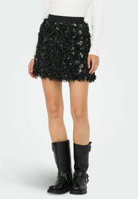 Zwarte minirok van gestructureerde stof, voorzien van pailletten en een zwarte elastische tailleband. Draagt zwarte leren kniehoge laarzen.