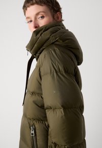 Chaqueta acolchada verde oliva con un cuello alto, bolsillos con cremallera y detalles cosidos. Presenta una textura brillante y suave, además de un diseño moderno.