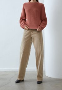 Pullover in Rostfarbe aus grob gestricktem Material mit rundem Halsausschnitt und Rippstruktur, kombiniert mit beigen weit geschnittenen Hosen und schwarzen Schuhen.