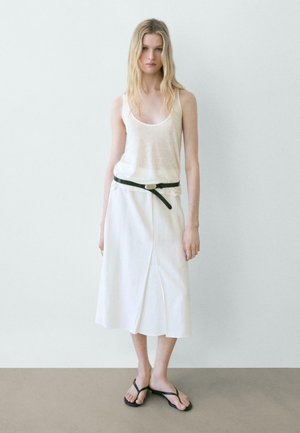 Femme aux longs cheveux blonds portant un haut blanc transparent sans manches, une jupe midi blanche avec une ceinture noire et des sandales noires, debout contre un mur uni.