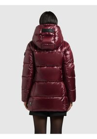 Nainen, joka pukeutunut kiiltävään viininpunaiseen hupulliseen toppatakkiin, seisoo selin vaaleanharmaata taustaa vasten.