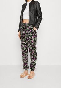 Pantalones de estampado floral negro con hojas verdes y flores moradas, con cintura y puños elásticos. Combinados con una chaqueta de cuero negra y sandalias.