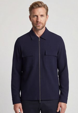 Man met licht haar die een marineblauwe jas met ritssluiting en twee borstzakken draagt, staand tegen een effen lichte achtergrond.