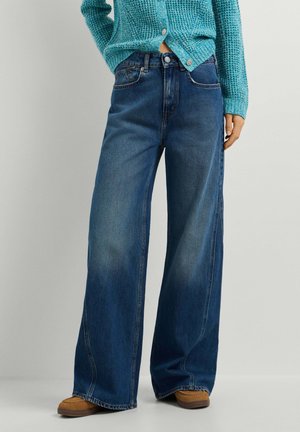 Frau trägt dunkelblaue Jeans mit weitem Bein, braune Schuhe und einen türkisen Strickpullover, steht vor einem schlichten hellen Hintergrund.