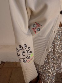 Pletený cardigan v smotanovej farbe s rebrovanými manžetami. Má vyšívané slnko a geometrické vzory v čiernej, červenej, modrej a neonovo žltej farbe.