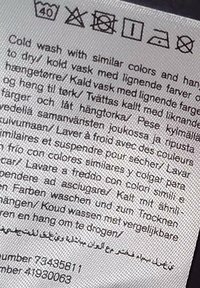 Vaskeetiket med instruktioner om kold vask ved 40°C, ingen blegning, ingen tørretumbling, lufttørring, lavt strygeniveau og ingen rensning med multilingual vejledning.
