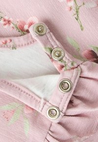 Tissu floral rose avec deux boutons-pression en métal, partiellement ouverts, montrant la doublure intérieure blanche du vêtement.