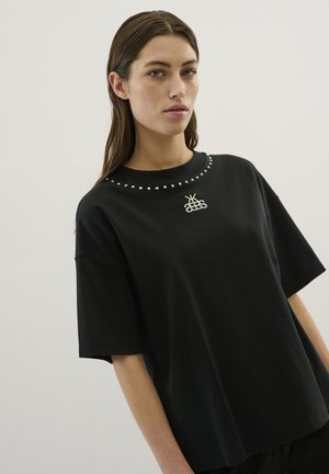 Sort bomulds t-shirt med rund hals, korte ærmer, sølvnitter rundt om kraven og hvidt broderet logo på forsiden.