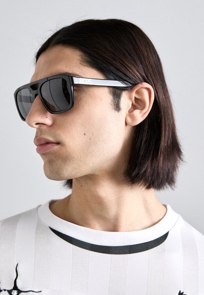 Gafas de sol negras de gran tamaño con lentes grises, con un marco rectangular audaz y un brazo con marca. Usadas por una persona que lleva una camisa blanca a rayas.