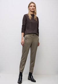 Donkerbruine trui met lange mouwen, gecombineerd met licht olijfgroene tapered broek. Model draagt zwarte enkel laarzen. Glad textuur, minimalistisch ontwerp.