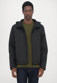 Schwarze Steppjacke mit Reißverschluss und Kapuze, mit matter Oberfläche und zwei vorderen Taschen, getragen über einem olivgrünen Rundhalsausschnitt-Shirt.