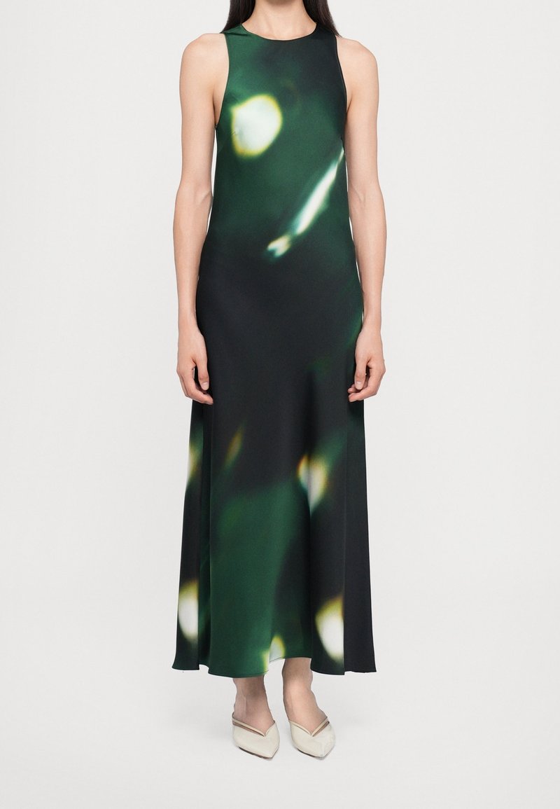 Robe longue sans manches avec un motif abstrait vert foncé et noir et des chaussures blanches pointues sur une femme debout.