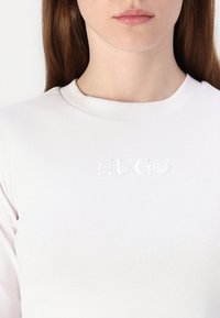 Valkoinen ribattuja pitkähihainen paita, jossa on kohokuvioitu "HUGO"-logo rinnassa. Pyöreä pääntie, jossa on sileä pinta.
